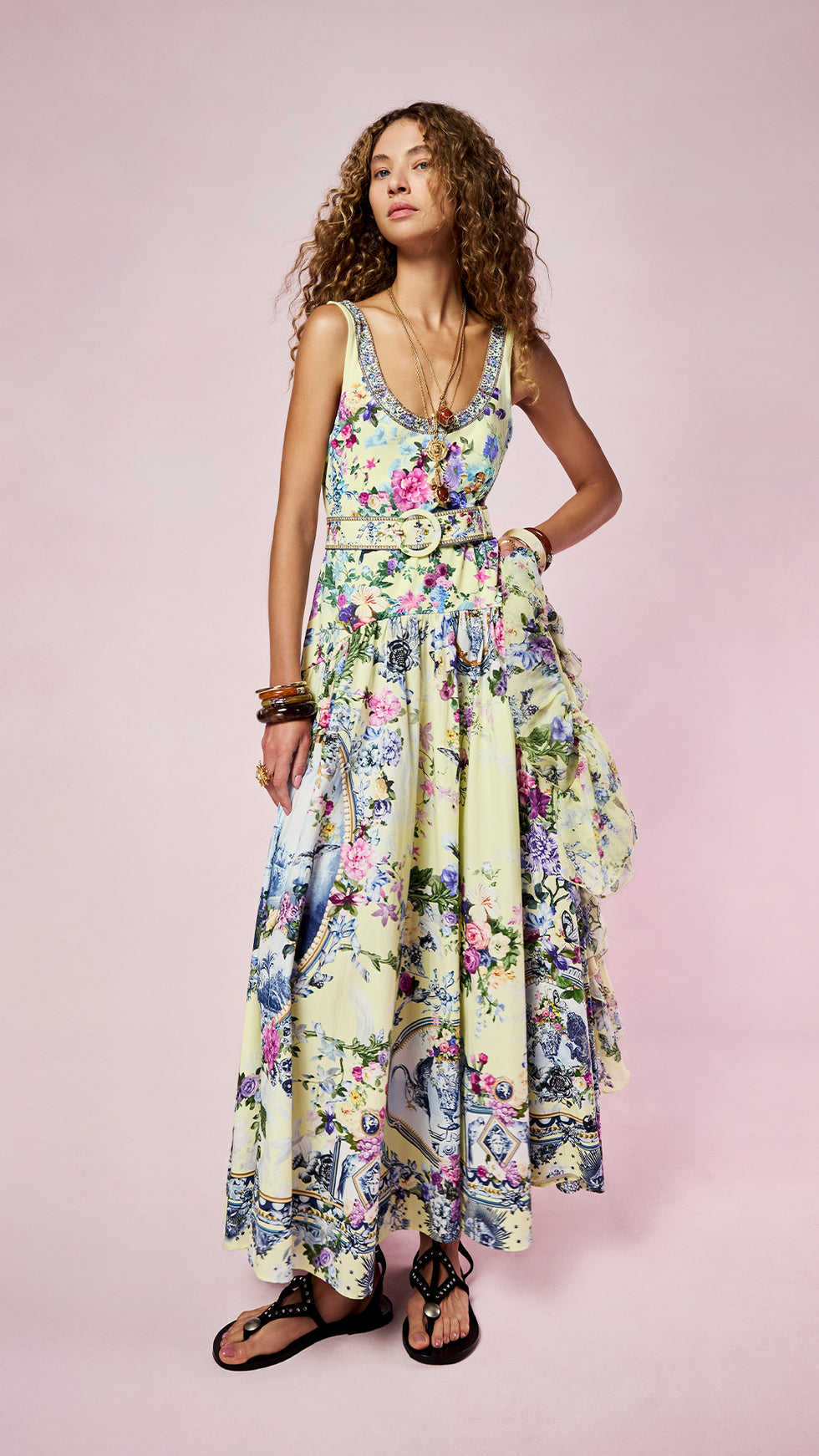 A woman modeling a sleeveless maxi dress with a colorful floral toile print on a pale yellow-green background, featuring a tie-waist belt and ruffled skirt, set against a solid pink studio backdrop.