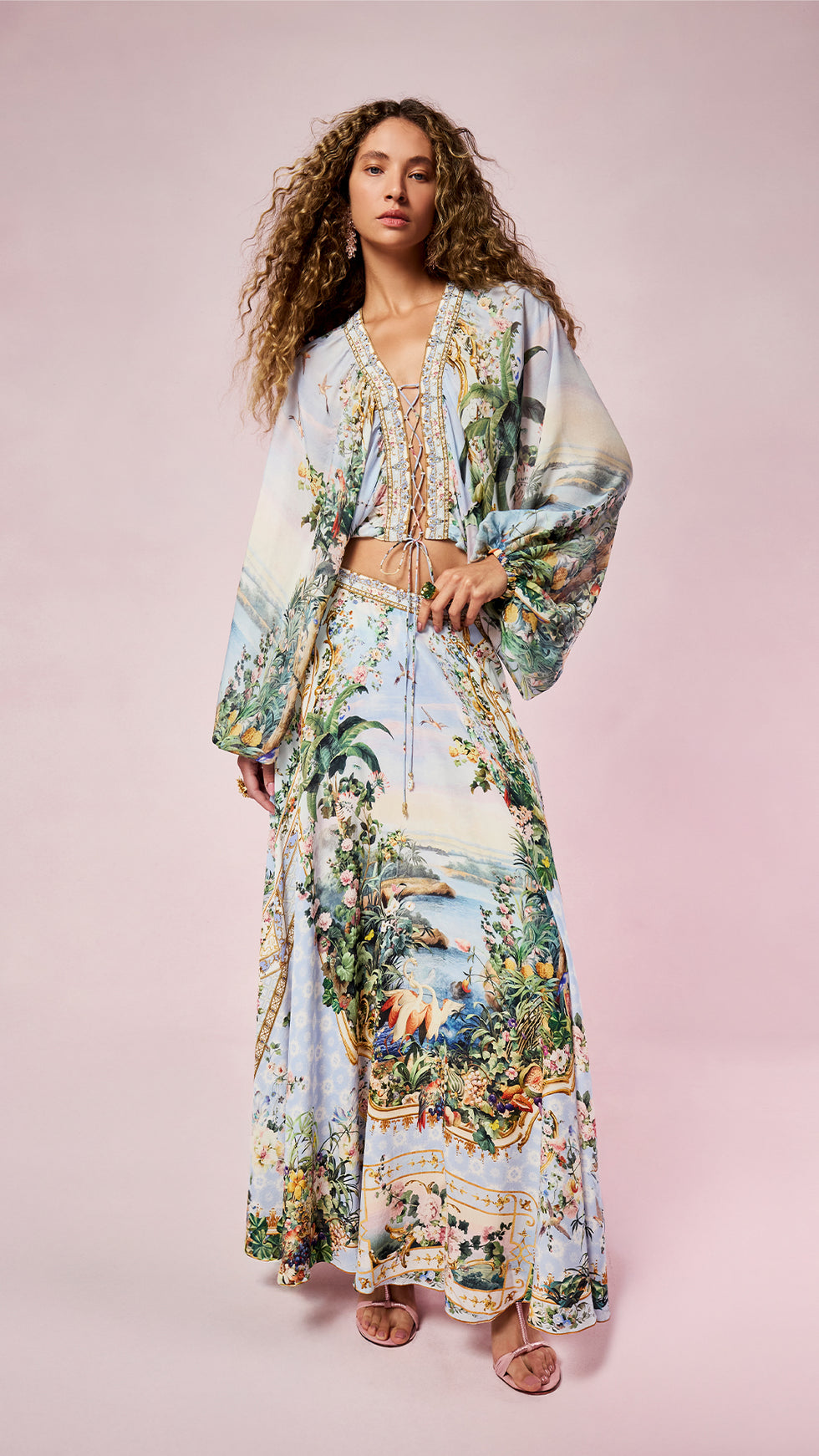 A woman modeling a matching two-piece bohemian maxi set with a detailed tropical landscape print, featuring a long-sleeve cropped tunic top and a long skirt, against a solid light pink background.